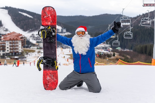 Um homem com um chapéu de Papai Noel com uma prancha de snowboard em uma estação de esqui.