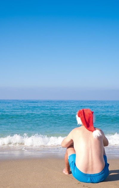 Um homem com um chapéu de Papai Noel sentado à beira-mar vista traseira Natal Papai Noel relaxando