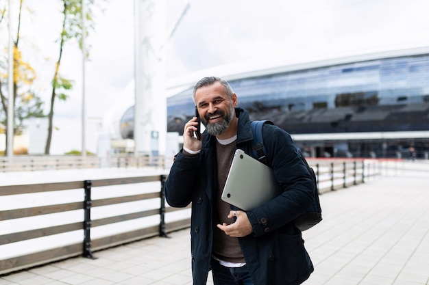 Um homem com um laptop nas mãos falando ao telefone andando pela rua