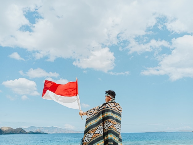 Um homem com um pano tradicional da indonésia chamado kain songket acenando com a bandeira da indonésia sob o céu azul