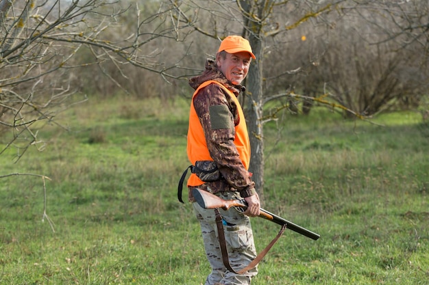 Um homem com uma arma nas mãos e um colete laranja em uma caça ao faisão em uma área arborizada em tempo nublado Caçador com cães em busca de caça