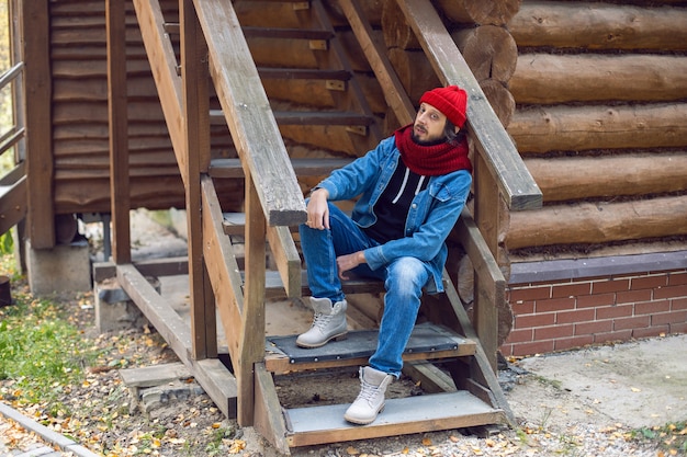 Um homem com uma barba hipster com um chapéu de malha vermelho e uma jaqueta jeans está sentado na escada