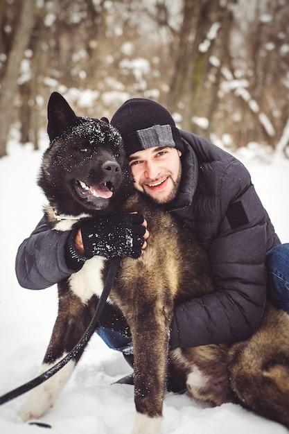 Um homem com uma jaqueta e um chapéu de tricô caminha por uma floresta nevada com um cachorro Akita americano