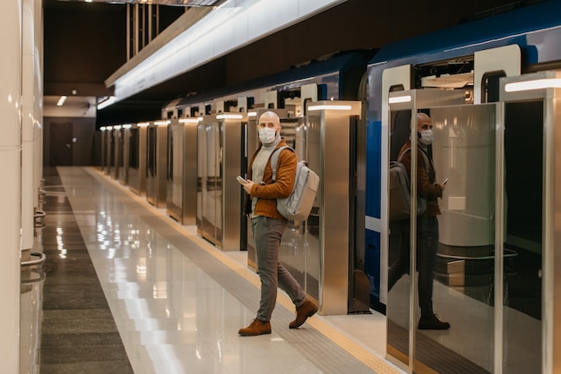 Um homem com uma máscara médica para evitar a disseminação do coronavírus está segurando um telefone celular enquanto sai de um moderno vagão do metrô. Um careca com máscara cirúrgica mantém distância social.