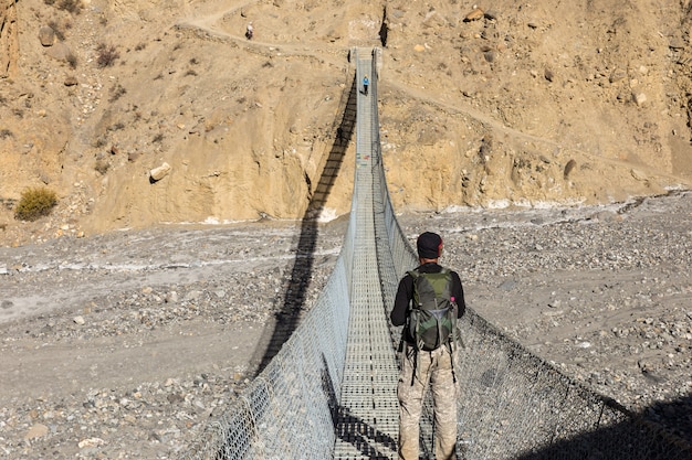 Um homem com uma mochila fica em uma ponte pedonal suspensa sobre um desfiladeiro da montanha e olha para as montanhas