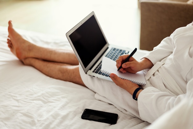 Um homem com uma túnica está deitado em uma cama com um computador nos pés, trabalho freelance de casa, sem rosto de um homem ...