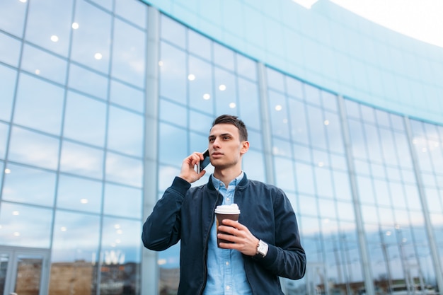 Um homem com uma xícara de café de papel atravessa a cidade, um cara bonito em roupas elegantes, ao telefone