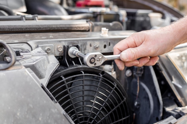 Um homem conserta um ventilador de refrigeração do motor sob a grelha do radiador de um carro Manutenção e reparo de máquinas