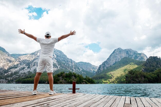 Um homem de branco fica contra o pano de fundo de um lago e montanhas com as mãos para cima uma bela paisagem vista traseira O conceito de liberdade felicidade sucesso vitória
