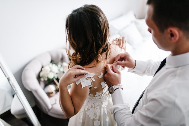 Um homem de camisa branca com gravata e relógio aperta os botões do espartilho do vestido. Noiva em vestido de noiva com renda em casa.