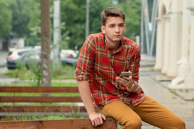 um homem de camisa xadrez senta-se em uma grade de madeira, segura um telefone nas mãos e digita uma mensagem