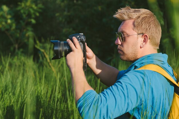 Um homem de camiseta azul e mochila amarela segura uma câmera fotográfica profissional nas mãos. Contra o pano de fundo da natureza verde e da floresta.