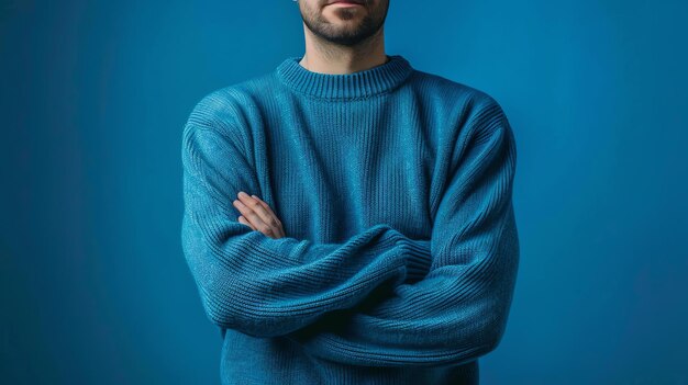 Um homem de camisola azul posa com os braços cruzados contra um fundo azul Ele tem barba e uma expressão séria no rosto