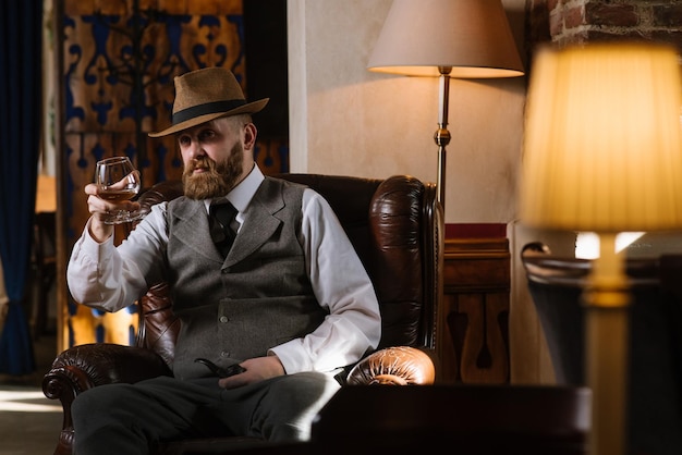 Um homem de chapéu vestindo terno vintage segurando cachimbo e copo de uísque sentado em uma cadeira grande