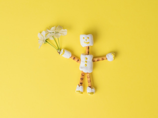 Um homem de marshmallow com um buquê de flores na mão sobre um fundo amarelo. Uma colagem de doces. Postura plana.