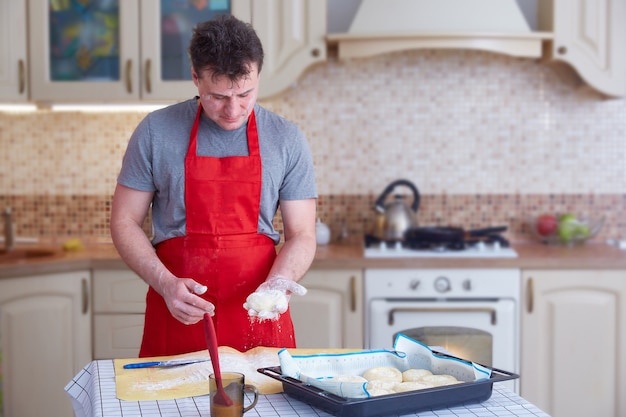 Um homem de meia-idade com um avental vermelho faz tortas de massa em casa na cozinha. Estilo de vida. Copie o espaço.