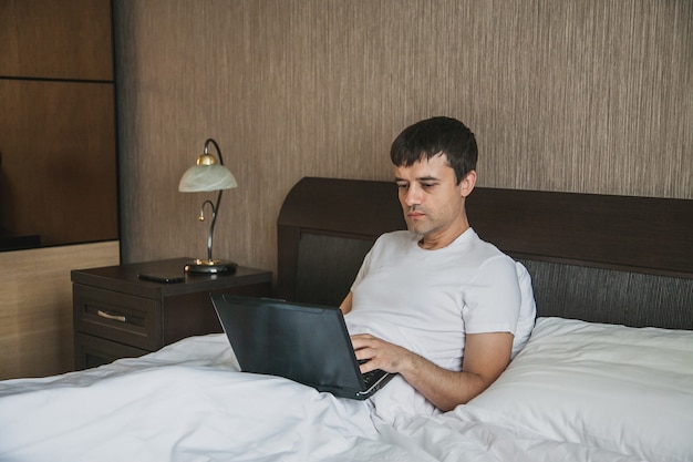 Um homem de meia-idade está sentado na cama em seu quarto e trabalhando em um laptop. O conceito de trabalho remoto