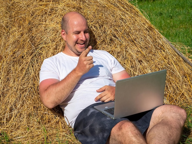 Um homem de meia idade vestido com uma camiseta e shorts está deitado em um palheiro amarelo com um laptop cinza nas mãos treinamento de trabalho remoto área rural expressão facial alegre emoção