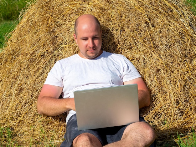 Um homem de meia idade vestido com uma camiseta e shorts está deitado em um palheiro amarelo com um laptop cinza nas mãos Treinamento de trabalho remoto Área rural