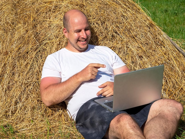 Um homem de meia-idade, vestido com uma camiseta e shorts, está deitado em um palheiro amarelo com um laptop nas mãos. Trabalho remoto, treinamento. Área rural. Um sorriso em seu rosto