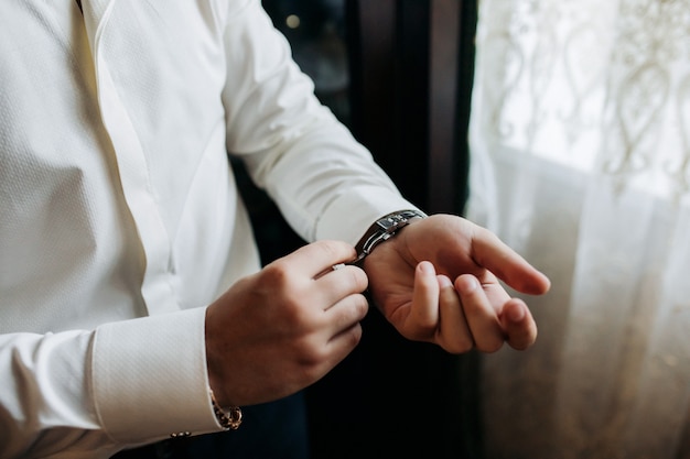 Um homem de negócios sexy prende um botão de punho no relógio de pulso, o punho da manga de uma camisa branca luxuosa.
