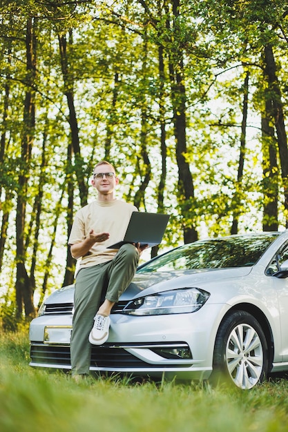 Um homem de óculos de sucesso trabalha em um laptop enquanto se apoia no capô de seu carro Trabalho remoto na natureza Trabalha em um laptop on-line Trabalha em um computador enquanto viaja