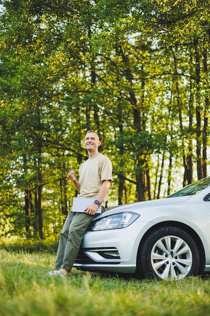 Um homem de óculos de sucesso trabalha em um laptop enquanto se apoia no capô de seu carro Trabalho remoto na natureza Trabalha em um laptop on-line Trabalha em um computador enquanto viaja