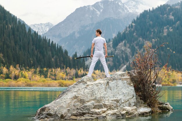 Um homem de roupas brancas com uma espada está de pé em uma grande pedra entre as terras altas.