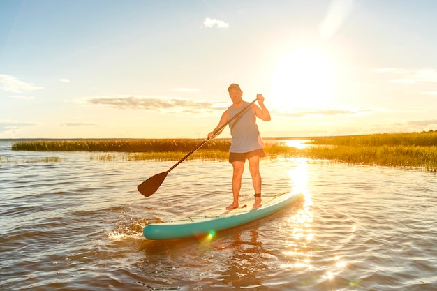 Um homem de shorts e uma camiseta em uma prancha de SUP com um remo flutua na água no contexto do pôr do sol