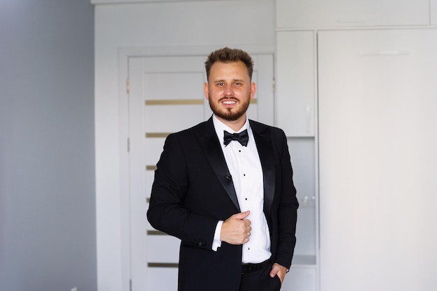 Um homem de smoking fica em frente a um armário branco com uma porta branca que diz "o noivo está vestindo um smoking preto"