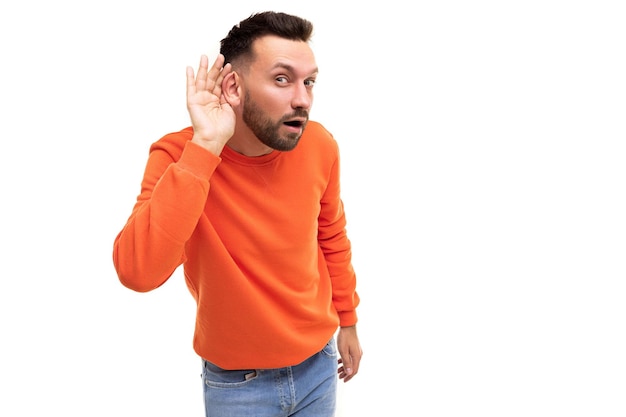 Um homem de suéter laranja ouve com a mão no ouvido em um fundo branco