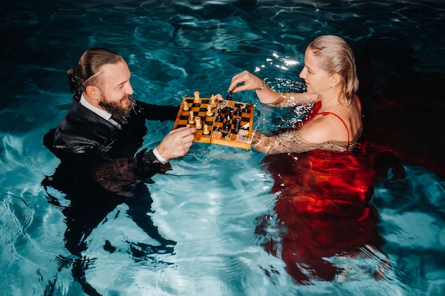 Um homem de terno e uma garota de vestido vermelho jogam xadrez na água da piscina.