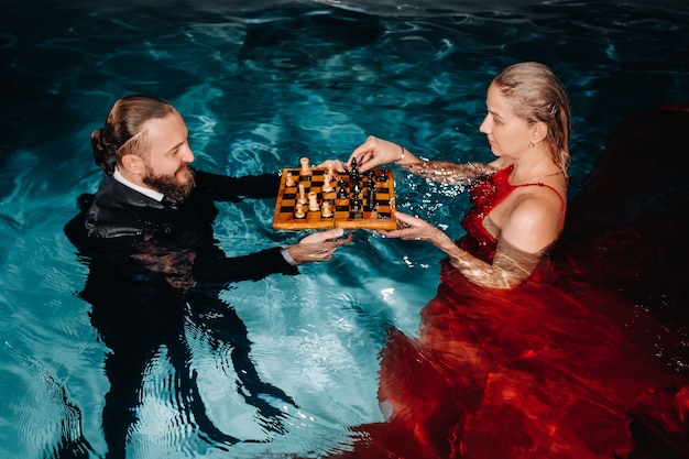 Um homem de terno e uma garota de vestido vermelho jogam xadrez na água da piscina
