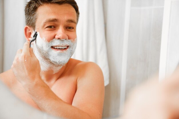 Um homem depila o rosto com um aparelho de barbear e se olha em um espelho redondo
