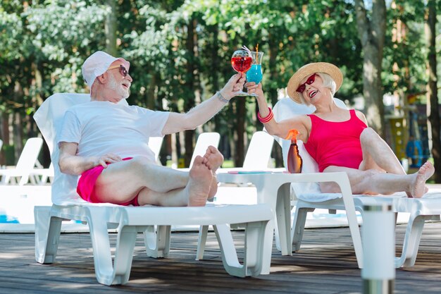 Um homem e uma mulher aposentados relaxando tomando coquetéis de verão enquanto tomam banho de sol perto da piscina