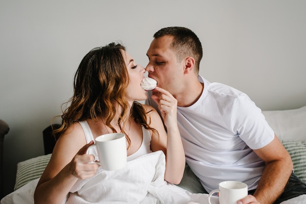 Um homem e uma mulher bebem café e comem marshmallows na cama