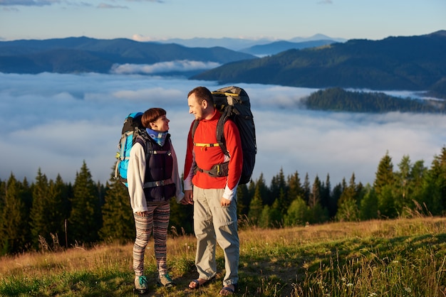 Um homem e uma mulher com mochilas se olham nos raios de um pôr do sol contra o pano de fundo da bela paisagem da floresta, montanhas e névoa sobre eles