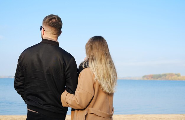 Um homem e uma mulher de casaco ficam de costas para a câmera e admiram a vista do lago