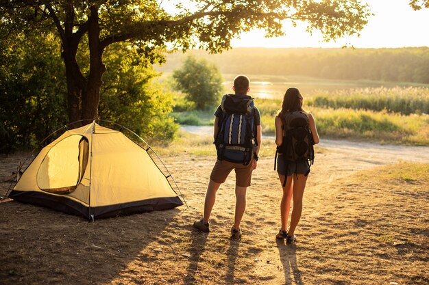 Um homem e uma mulher em uma caminhada com mochilas perto de uma tenda ao pôr do sol. lua de mel na natureza