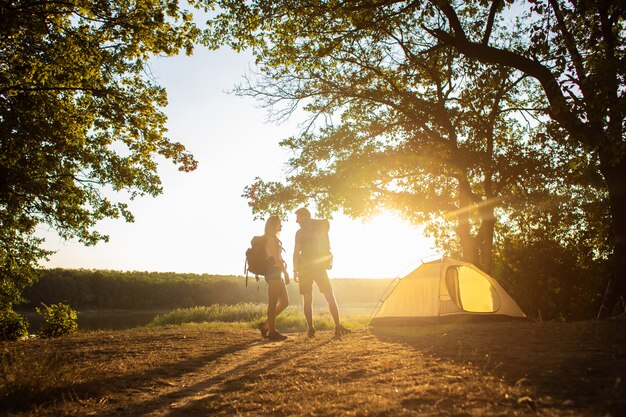 Um homem e uma mulher em uma caminhada com mochilas perto de uma tenda ao pôr do sol. Lua de mel na natureza