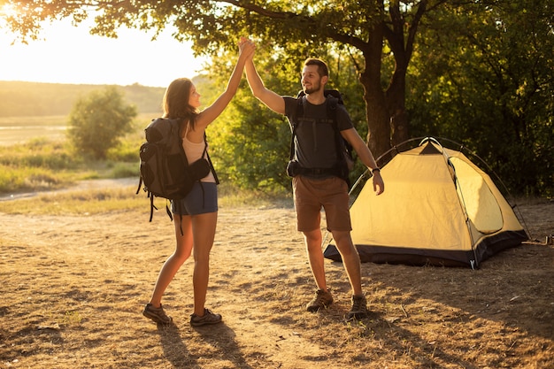 Um homem e uma mulher em uma caminhada com mochilas perto de uma tenda ao pôr do sol, olá cinco. Lua de mel na natureza