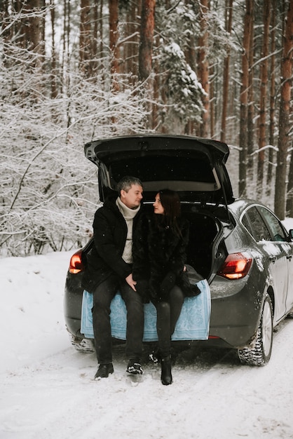 Um homem e uma mulher no fundo de uma floresta coberta de neve em uma nevasca no porta-malas de um carro