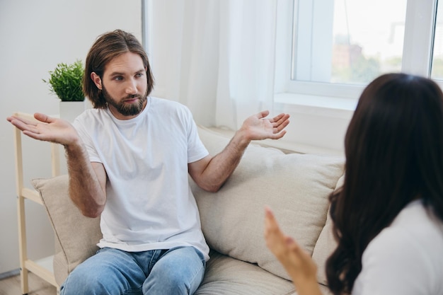 Um homem e uma mulher sentam-se no sofá em casa em camisetas brancas e conversando escandalosamente não se entendem Uma briga na família de dois cônjuges e agressão