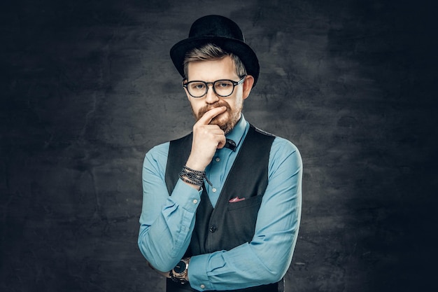 Um homem elegante hipster barbudo vestido com uma camisa azul, colete e chapéu de cilindro sobre fundo cinza vinheta.