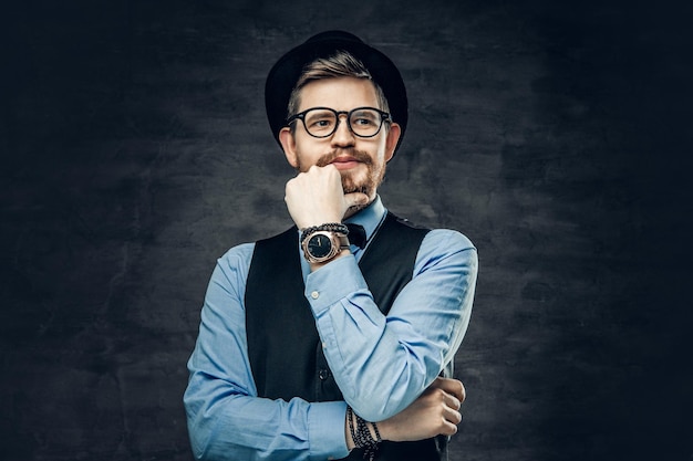 Um homem elegante hipster barbudo vestido com uma camisa azul, colete e chapéu de cilindro sobre fundo cinza vinheta.