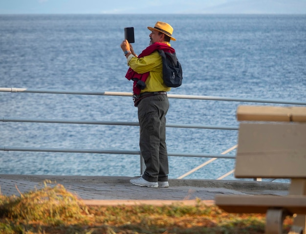 Um homem elegantemente vestido com um chapéu tira uma foto do mar no aterro na Grécia