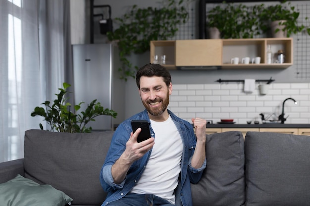 Um homem em casa ganhou e enfrenta as notícias online olha para a tela do telefone e comemora o triunfo da vitória freelancer com telefone