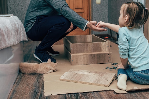um homem em casa recolhe móveis novos, uma filhinha observa seu trabalho e ajuda
