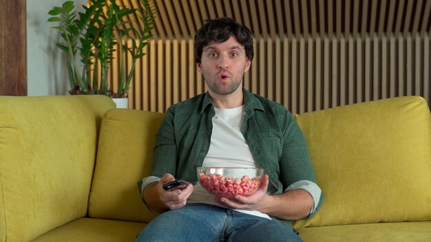 Um homem em frente à tv assistindo a um jogo de esportes ou um filme, sentado em um sofá amarelo e comendo pipoca