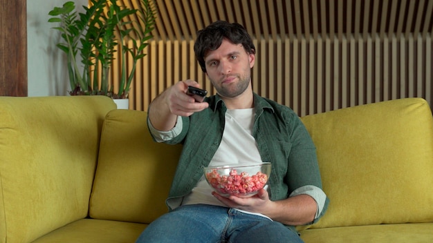 Foto um homem em frente à tv assistindo a um jogo de esportes ou um filme, sentado em um sofá amarelo e comendo pipoca